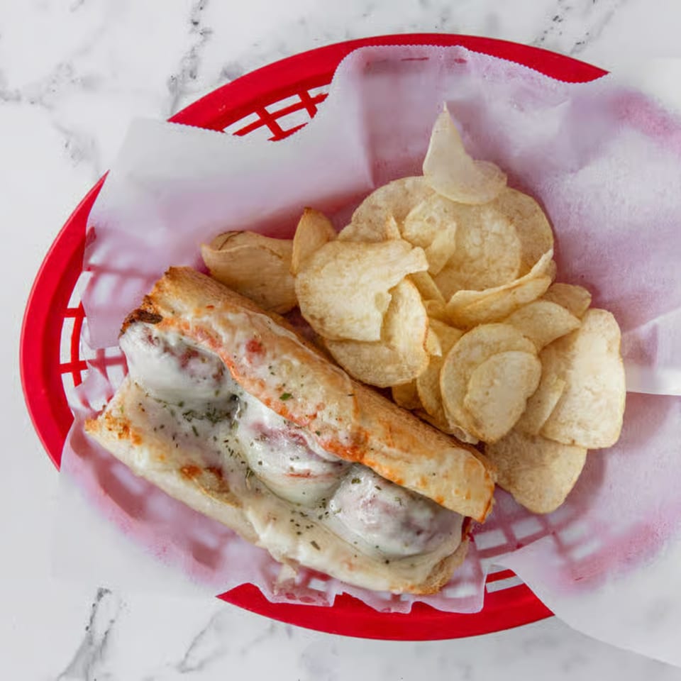 Meatball sub sandwich with melted cheese and potato chips in a red basket.